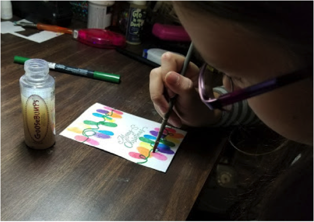 A girl applies GooseBumps texture medium to her handmade card featuring a string of Christmas lights.